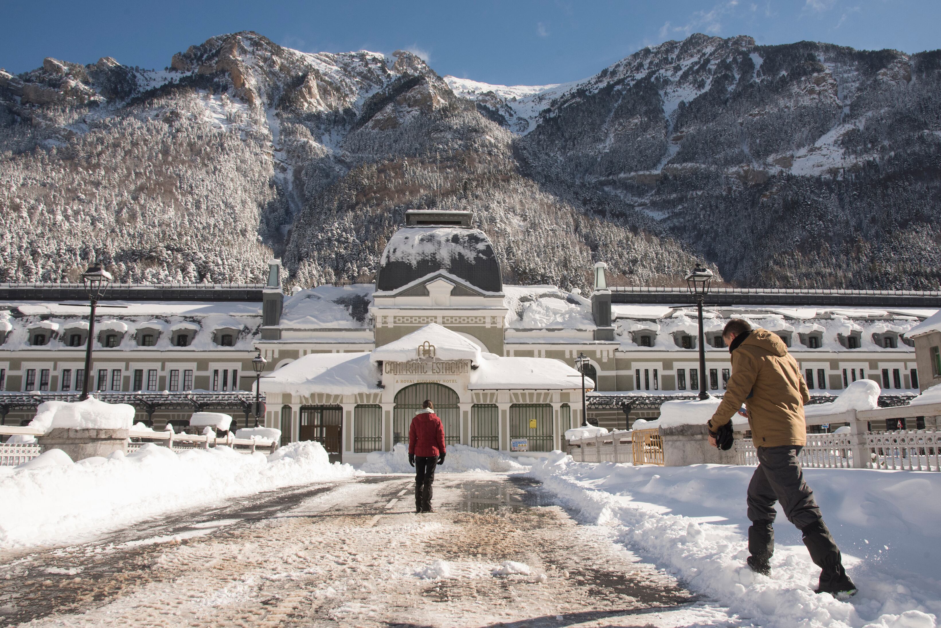 Varias personas pasan delante del edificio de la antigua estación de tren de Canfranc (Huesca), cubierta por la nieve acumulada.