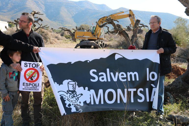 Dos membres de la plataforma Salvem lo Montsià sostenen la pancarta i algun cartell contra l&#039;espoli d&#039;oliveres davant una retroexcavadora que arrenca al fons els arbres de la finca de Cultius Ponç.