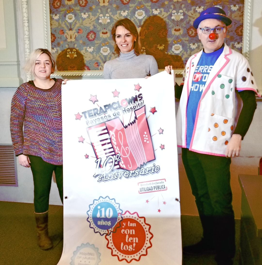 Alicia y Carlos (Terapiclowns) flanqueando a la concejala de cultura, Lorena de la Fuente, con el cartel del 10 aniversario de la asociación