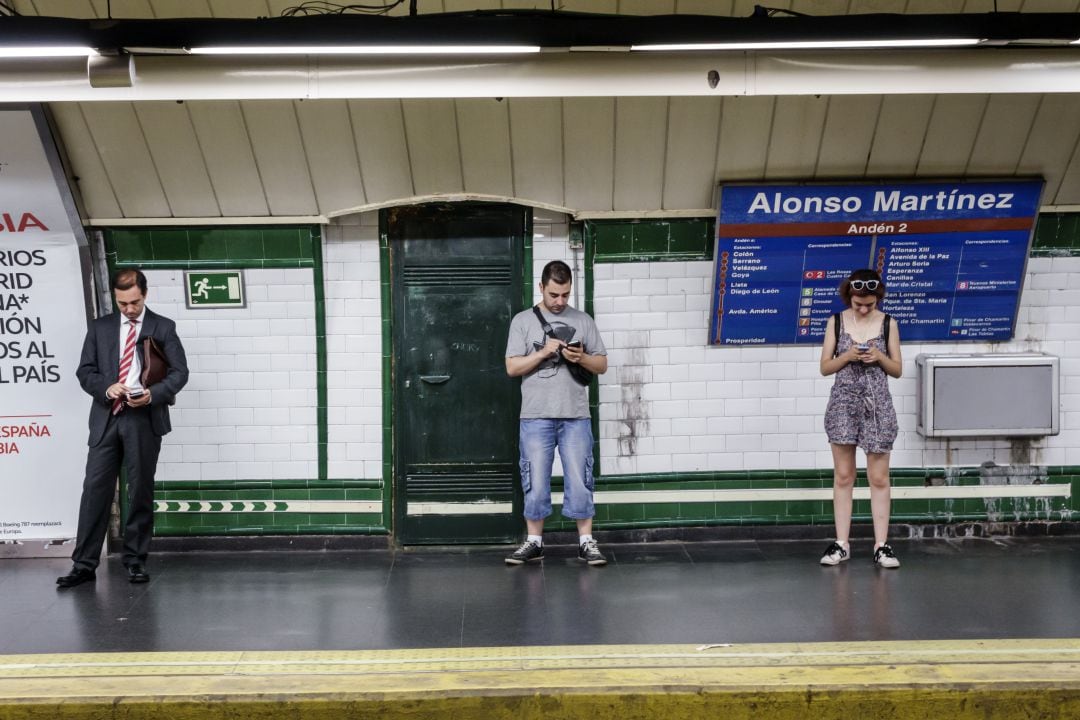 Varios ciudadanos miran el móvil mientras esperan la llegada del metro en la estación de Alonso Martínez