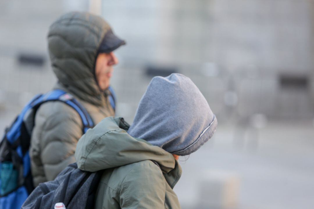 Dos jovenes se protegen del frío con abrigos y gorros mientras pasean durante un día de viento por Madrid (España).