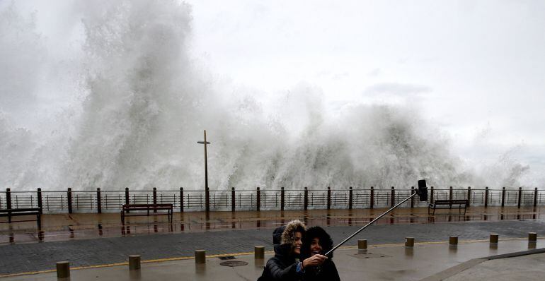 Dos jóvenes se fotografían con una ola rompiendo en el Paseo Nuevo de San Sebastián.