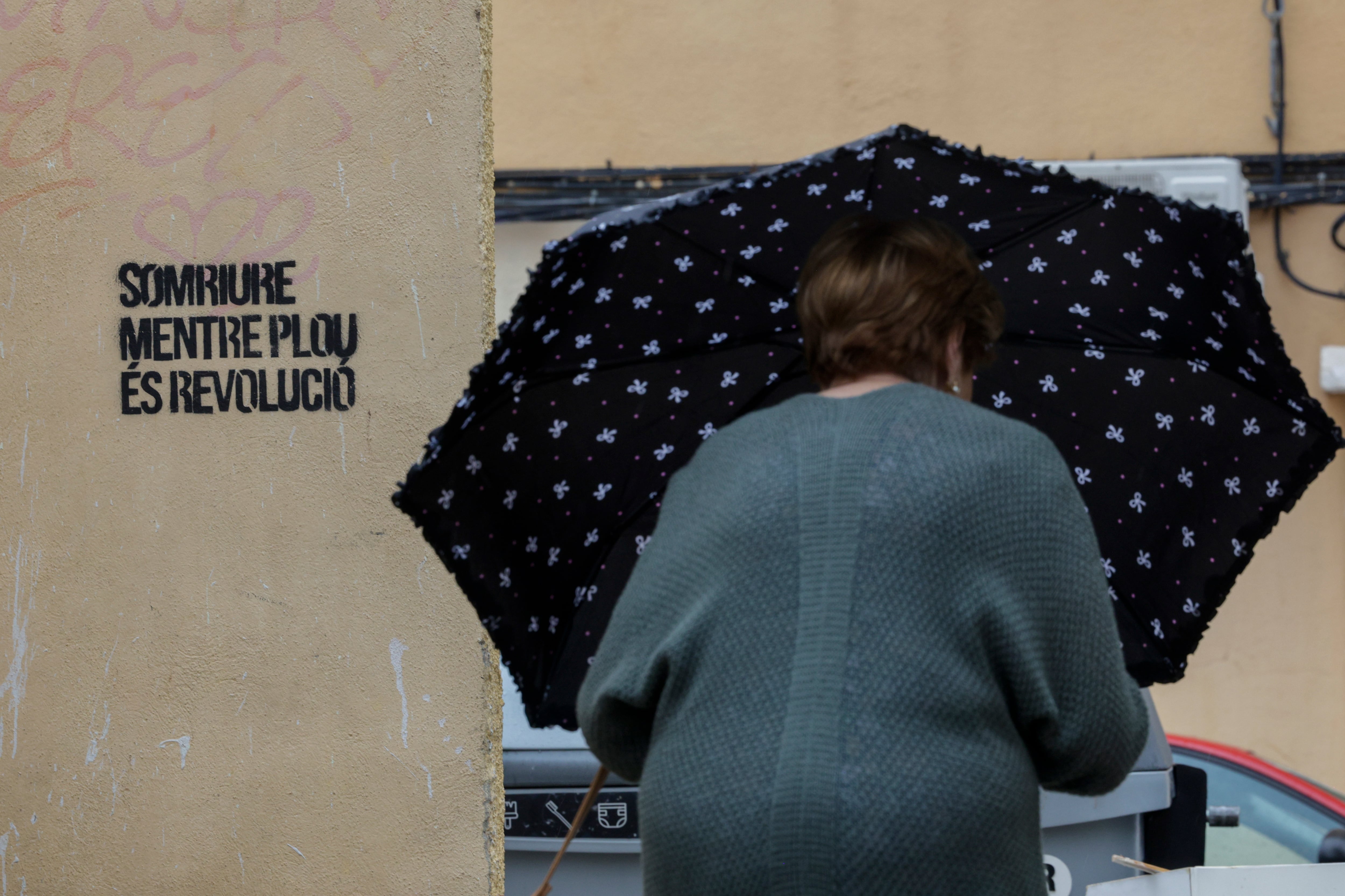 GRAFCVA9470. BURJASSOT (VALENCIA), 29/09/2025.- Una mujer se protege de la lluvia con un paraguas cuando pasa junto a una pintada con el lema "Sonreir mientras llueve es revolución" EFE/Kai Försterling