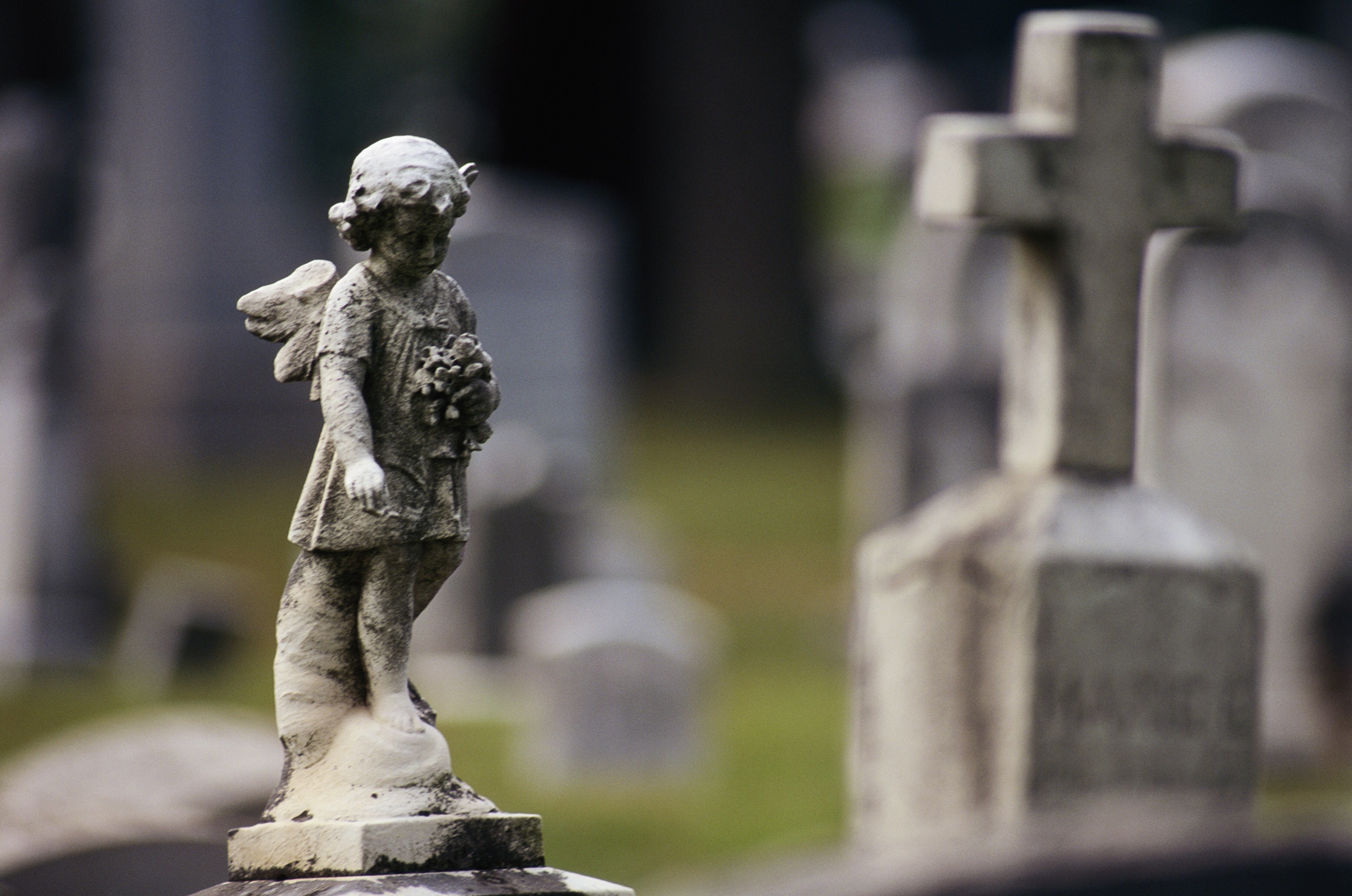 Escultura de un ángel, en primer plano, con tumbas de un cementerio de fondo