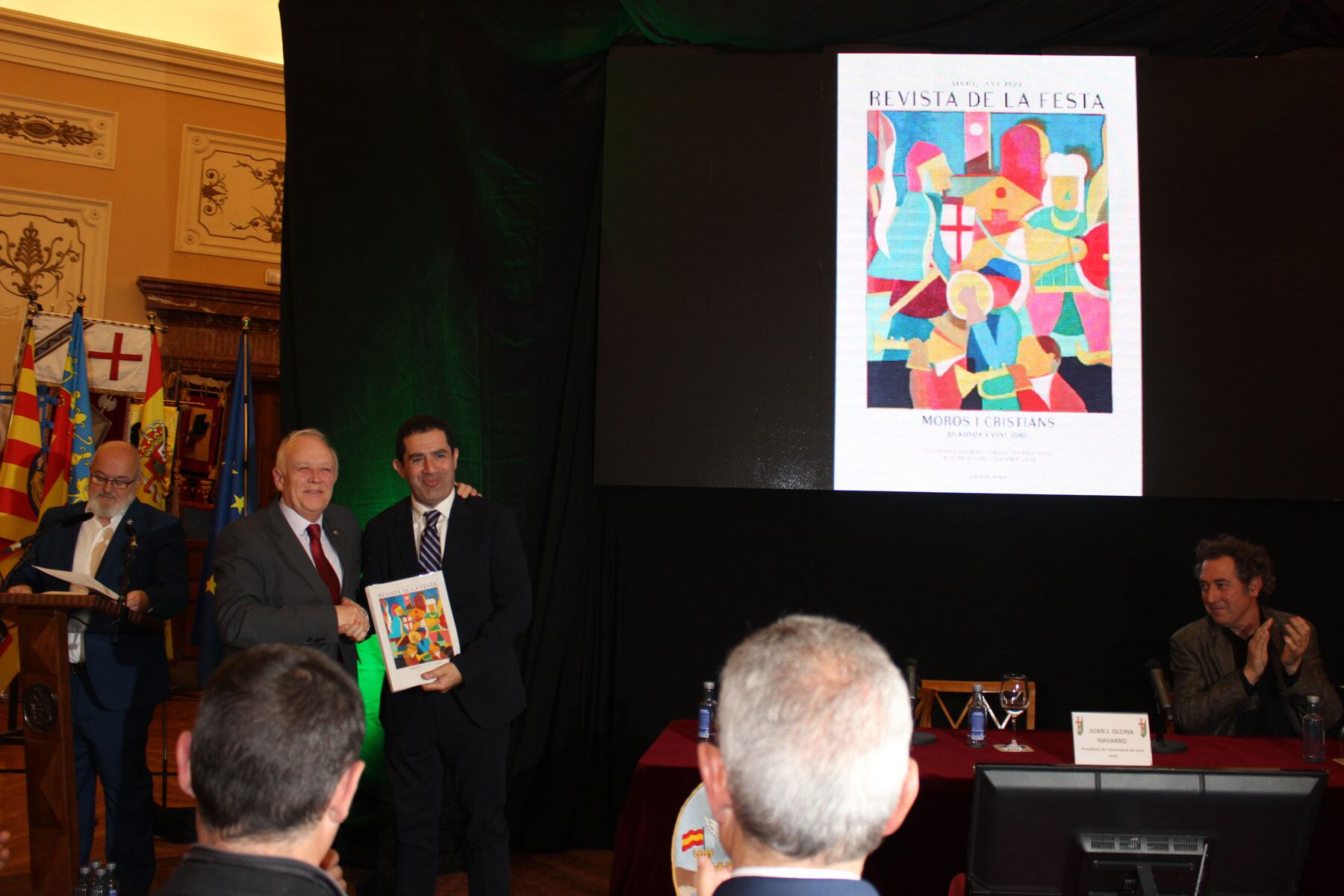 El presidente de la Asociación de San Jorge, Juan José Olcina, entrega el primer ejemplar de la revista al alcalde de Alcoy, Antonio Francés, con el autor de la portada, Jordi Vila, al fondo y la portada proyectada