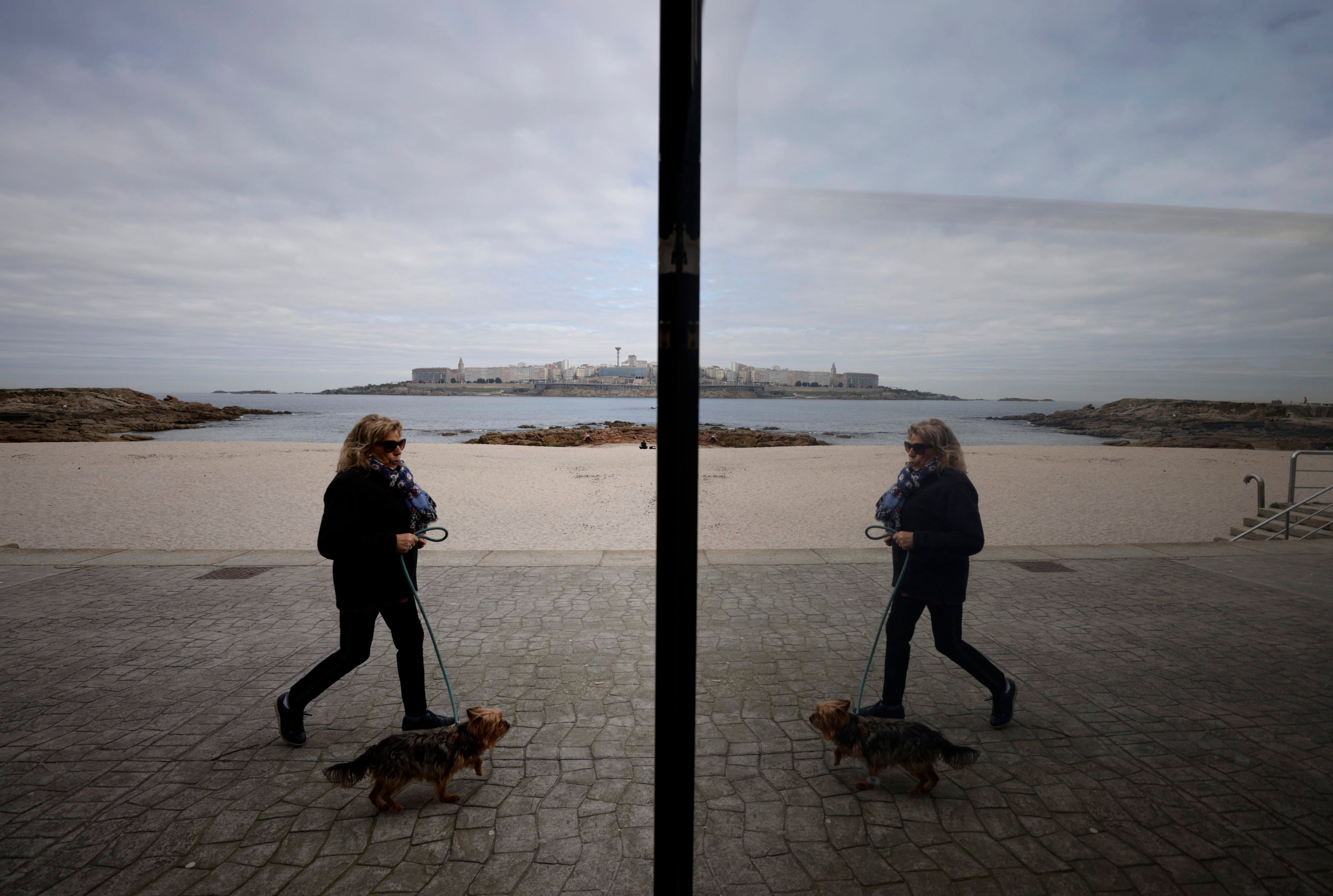 A CORUÑA, 06/03/2023.- Una mujer y su perro caminaban este lunes por el paseo marítimo de A Coruña, con su imagen reflejada en un cristal de un local de hostelería este lunes. El tiempo arranca esta semana con varios frentes que dejarán lluvias en el país, hasta el viernes, sobre todo en la mitad occidental, mientras las temperaturas subirán de forma clara dejando atrás el frío invernal de los últimos días para llegar al fin de semana con valores propios de mayo, e incluso de junio. EFE/Cabalar
