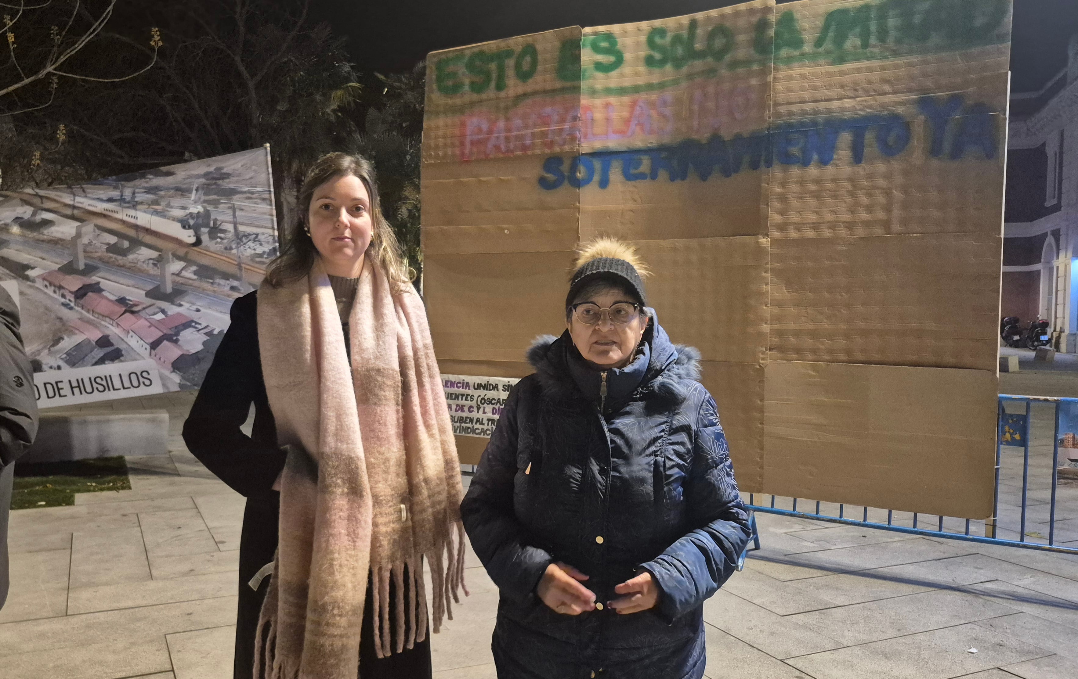 Cristina Martin Moro (i) y Maite Rodríguez (d), de la Federación de Asociaciones de Vecinos de Palencia, frente a la pantalla ficticia que han colocado frente a la estación como un acto reivindicativo y de protesta