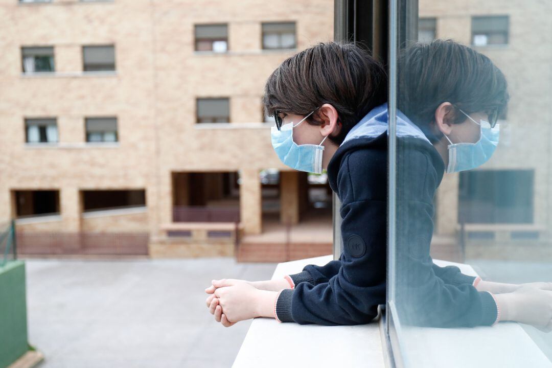 Un niño con una mascarilla se asoma a la ventana de su casa.