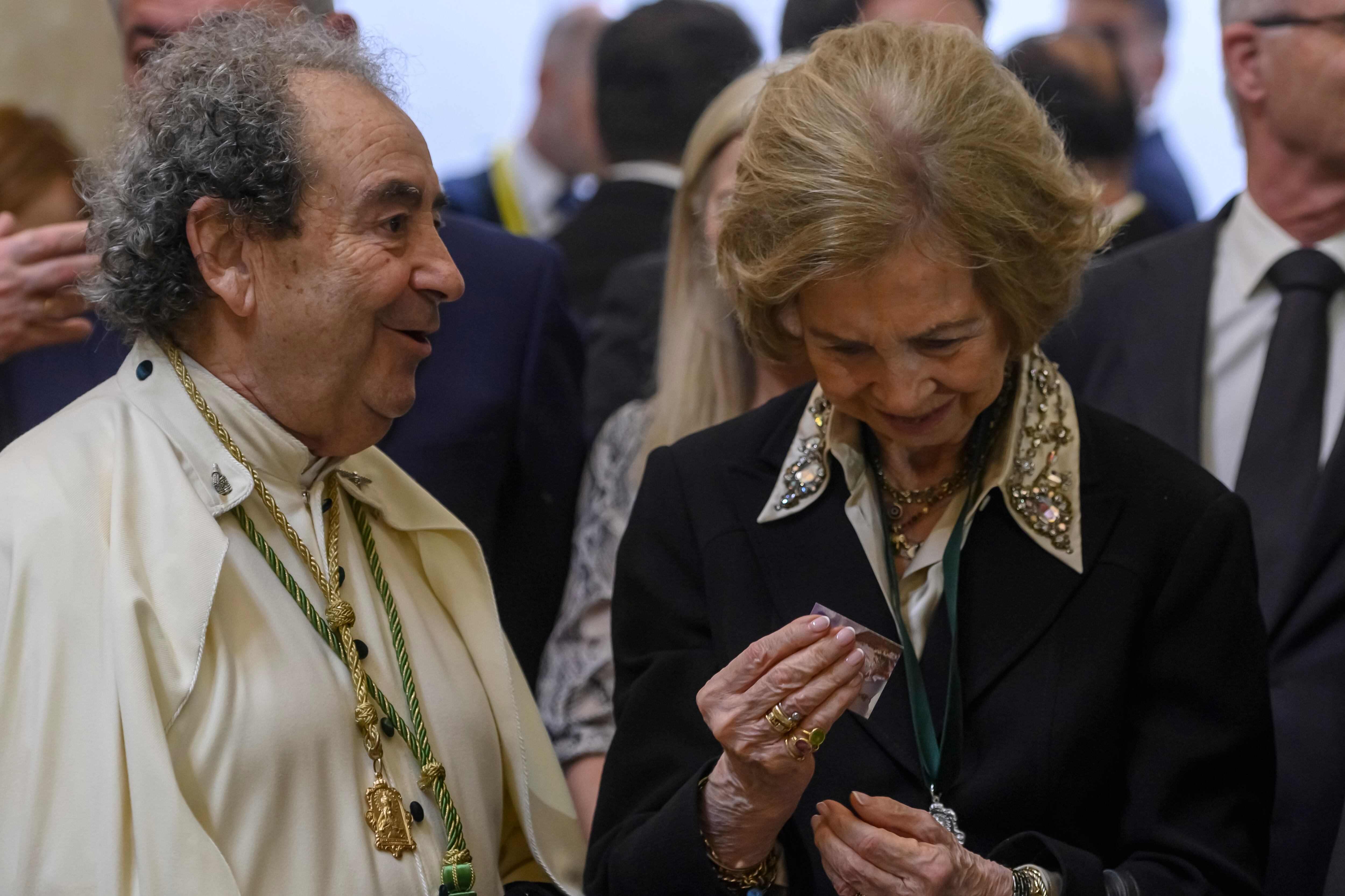 La Reina Sofía acompañada por el Hermano Mayor de la Hermandad de La Macarena, José Antonio Fernández (i), observa una estampa entregada por un nazareno durante la salida de la cofradía este jueves, en Sevilla (España). EFE/ Raúl Caro