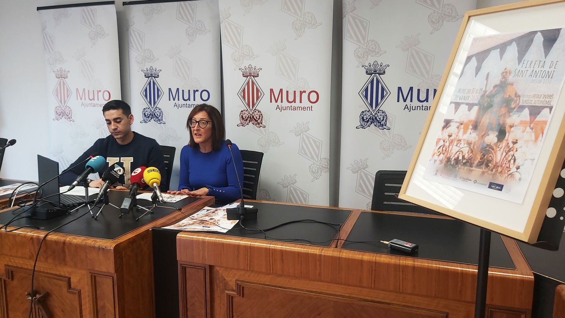 Carol Ivàñez i Fernando Picó, en el saló de plenaris de l&#039;Ajuntament de Muro amb un cartell de la Fireta de Sant Antoni al costat