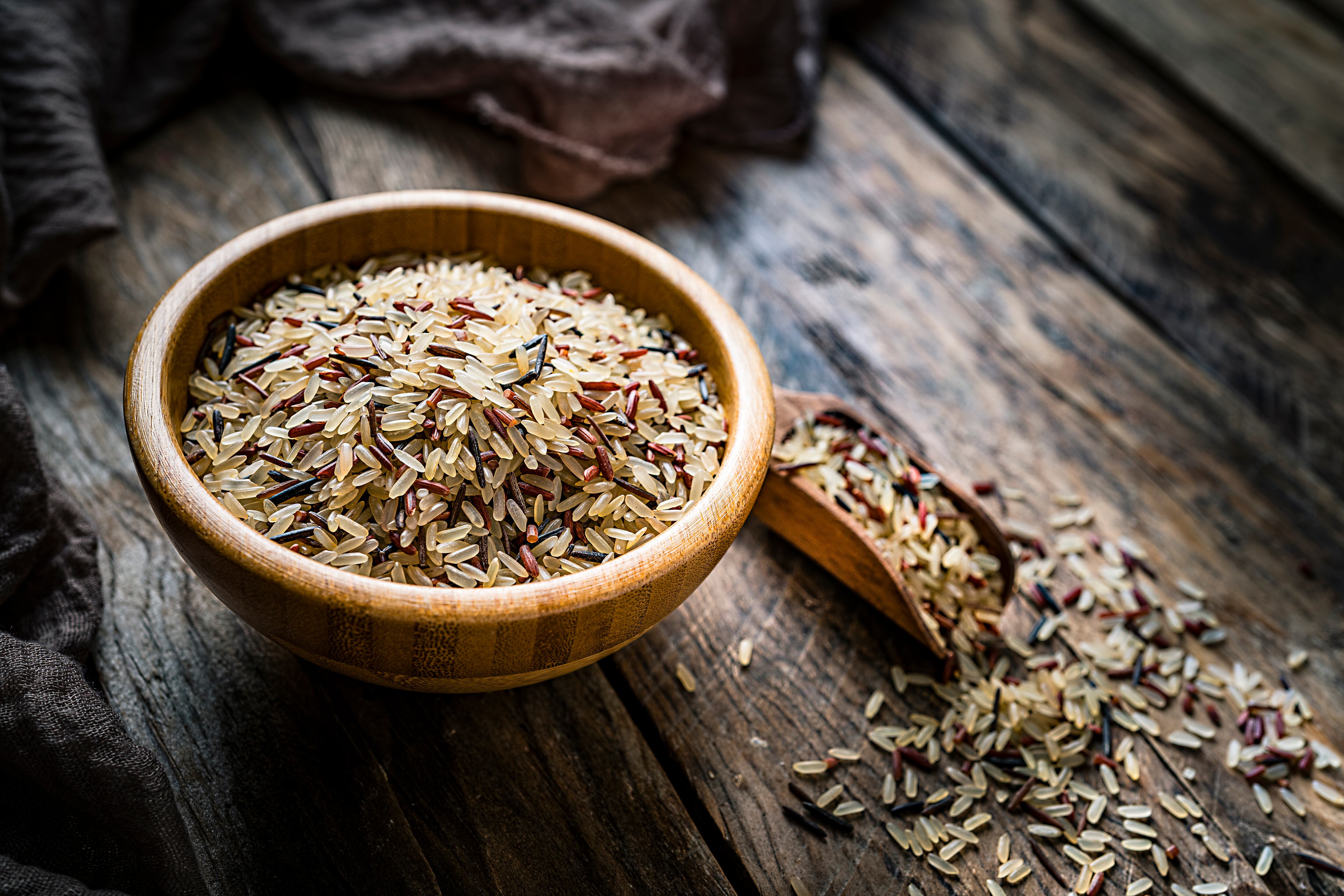 High angle view of a wooden bowl filled with mixed rice on a rustic wooden table. A wooden serving scoop is beside the bowl. Predominant color is brown. High resolution 42Mp studio digital capture taken with SONY A7rII and Zeiss Batis 40mm F2.0 CF lens