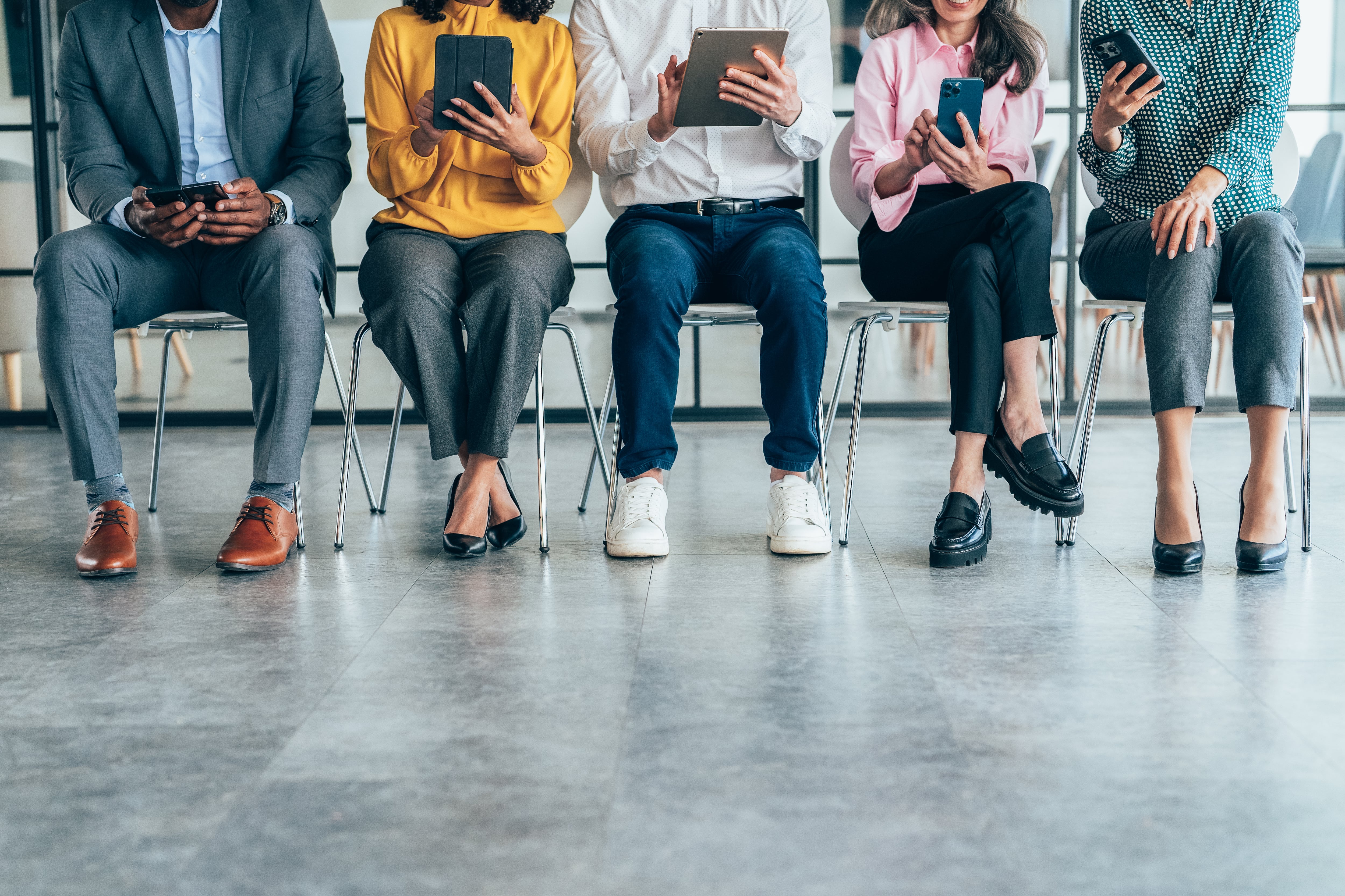 A row of individuals seated in chairs, each engaged with their smartphones or tablets. The image highlights modern technology usage and diversity in a casual setting, showcasing a mix of ages, styles, and devices. Business people, networking and social media at the office using technology at the workplace. Diverse group of creative workers in design for digital tech company marketing mobile communication
