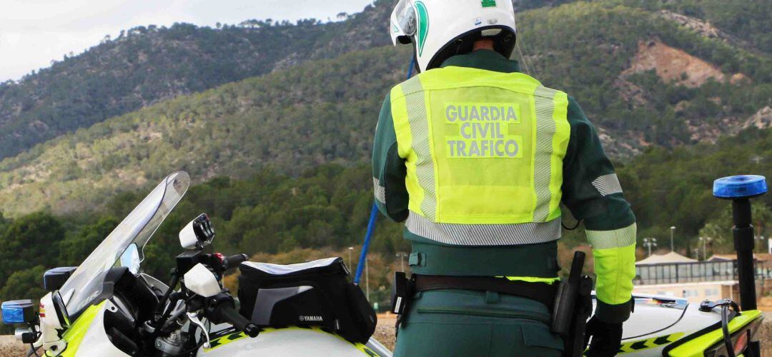 Un agente de la Guardia Civil de Tráfico, de espaldas, en una carretera junto a su moto y una montaña al fondo.