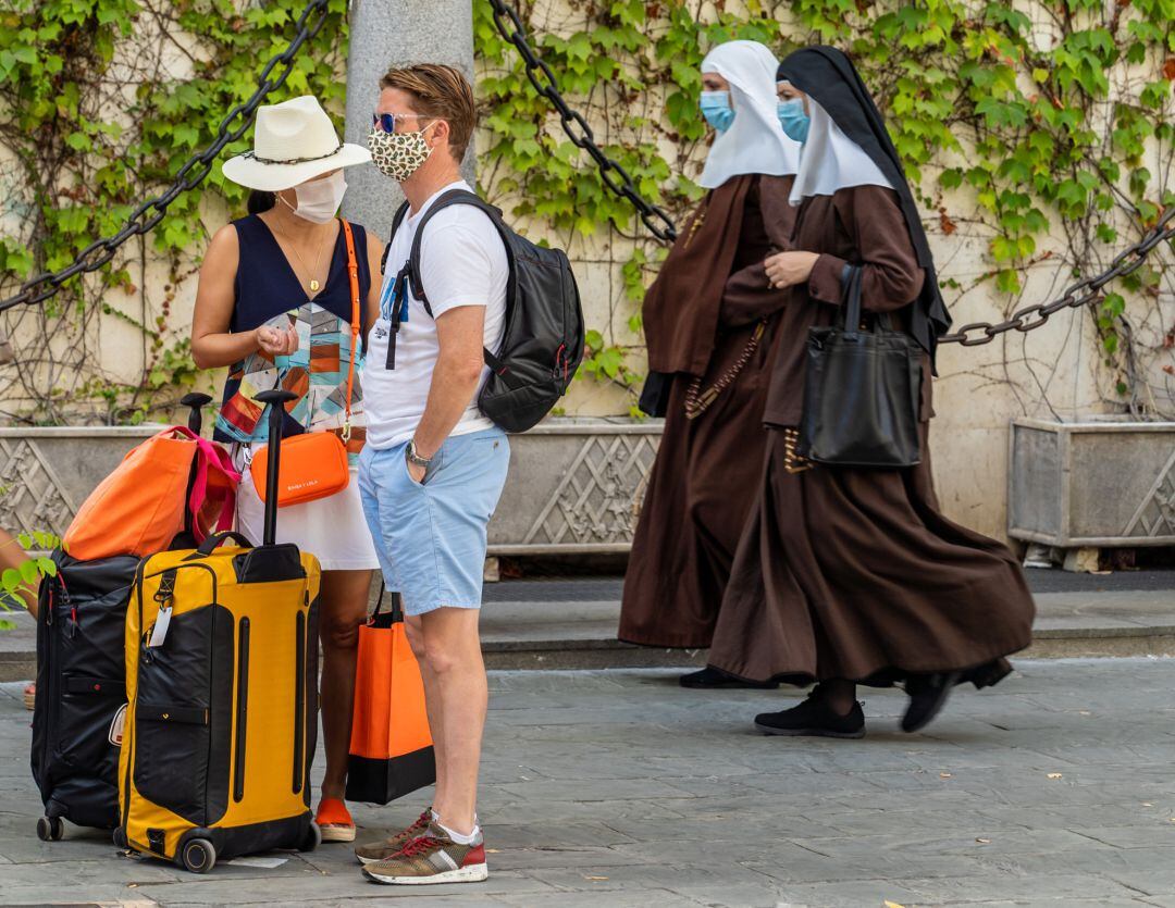 Unos turistas esperan el autobús mientras una pareja de monjas de la Orden de Santa Ángela de la Cruz  pasan por detrás con mascarillas.
