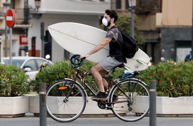 Un surfista protegido con mascariilla carga su tabla en bici hacia la playa de la Malvarrosa, en Valencia, hoy quincuagésimo sexto día del estado de alarma