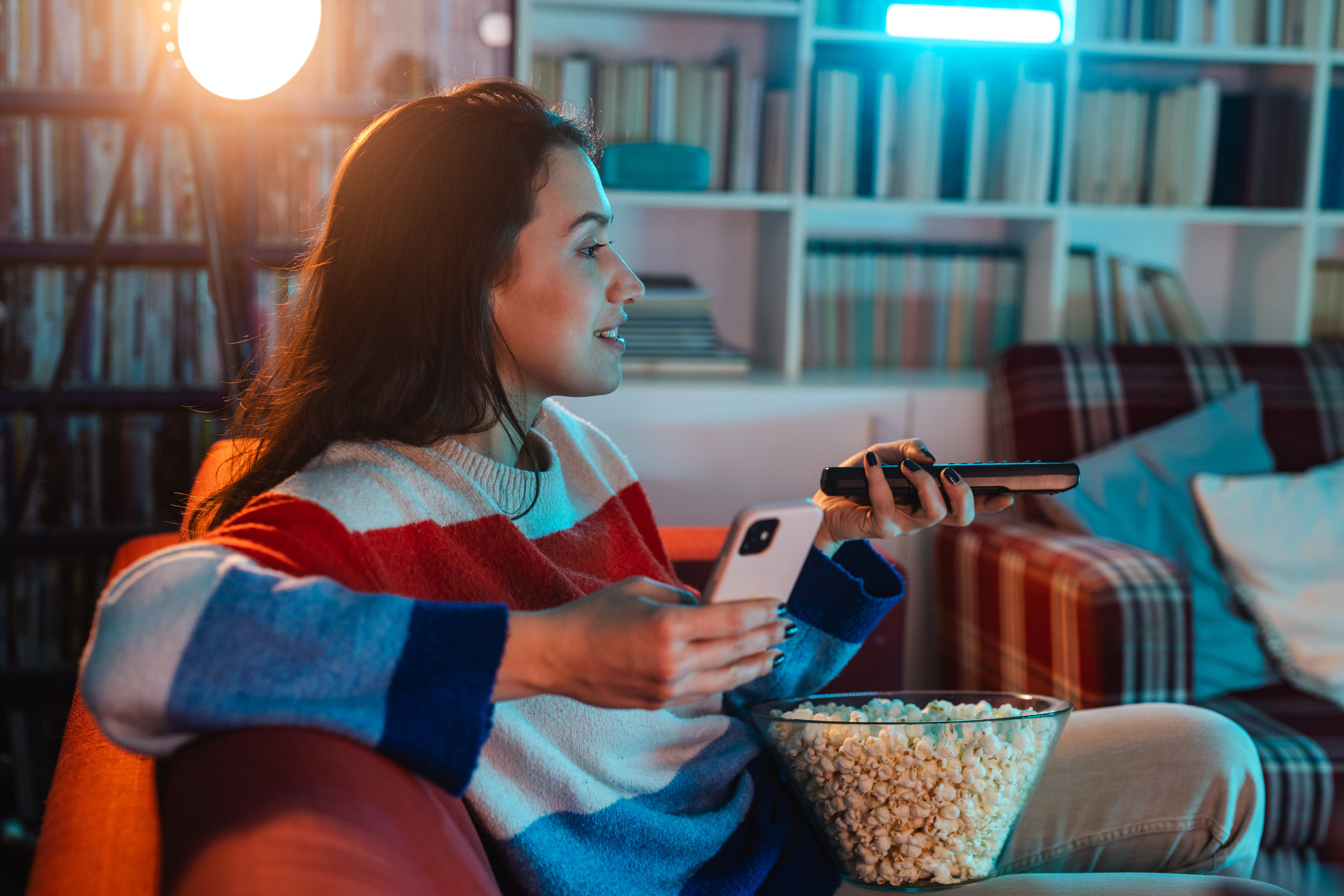 Young woman relaxing at home, eating popcorn and using phone while watching TV