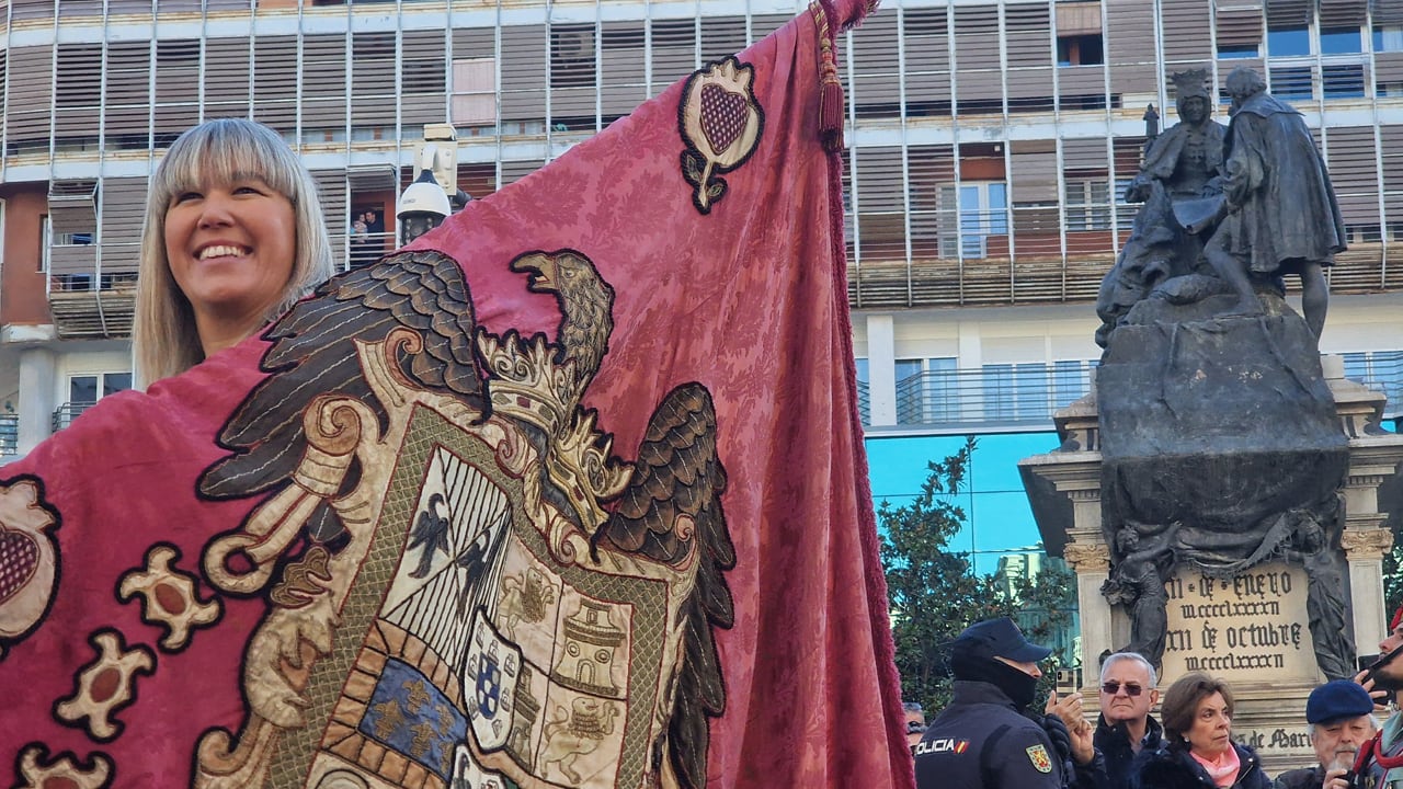 La concejala Carolina Amate porta el Pendón de Castilla, en la Gran Vía de Granada en la fiesta de la Toma de la ciudad del 2 de enero de 2024 con el monumento a las Capitulaciones de Santa Fe, entre Isabel la Católica y Cristóbal Colón, de fondo