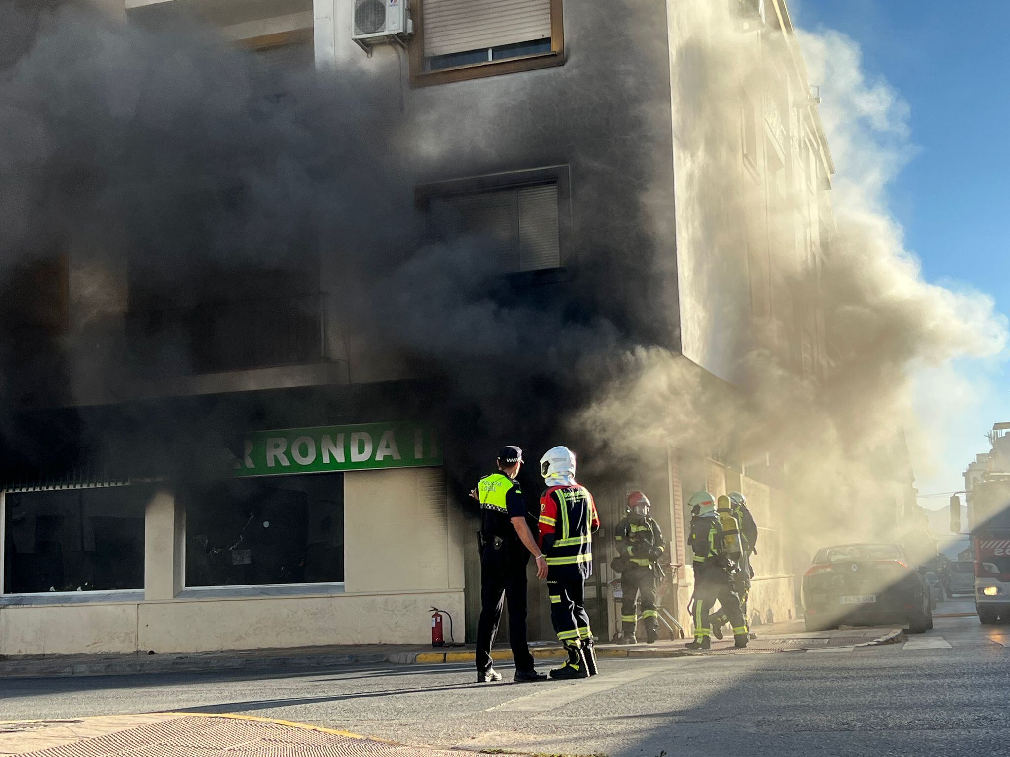 Varios bomberos junto a un efectivo de la Policía Local ante el edificio envuelto en humo