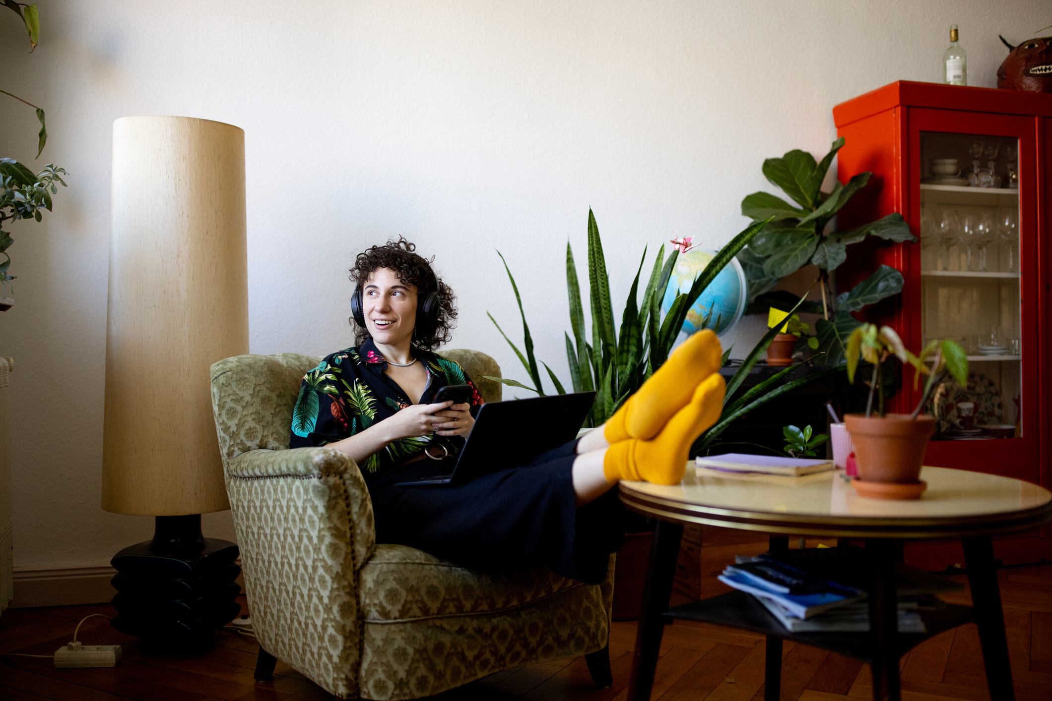 Non-binary person wearing headphones listening to music from her mobile phone while sitting with a laptop on the armchair.