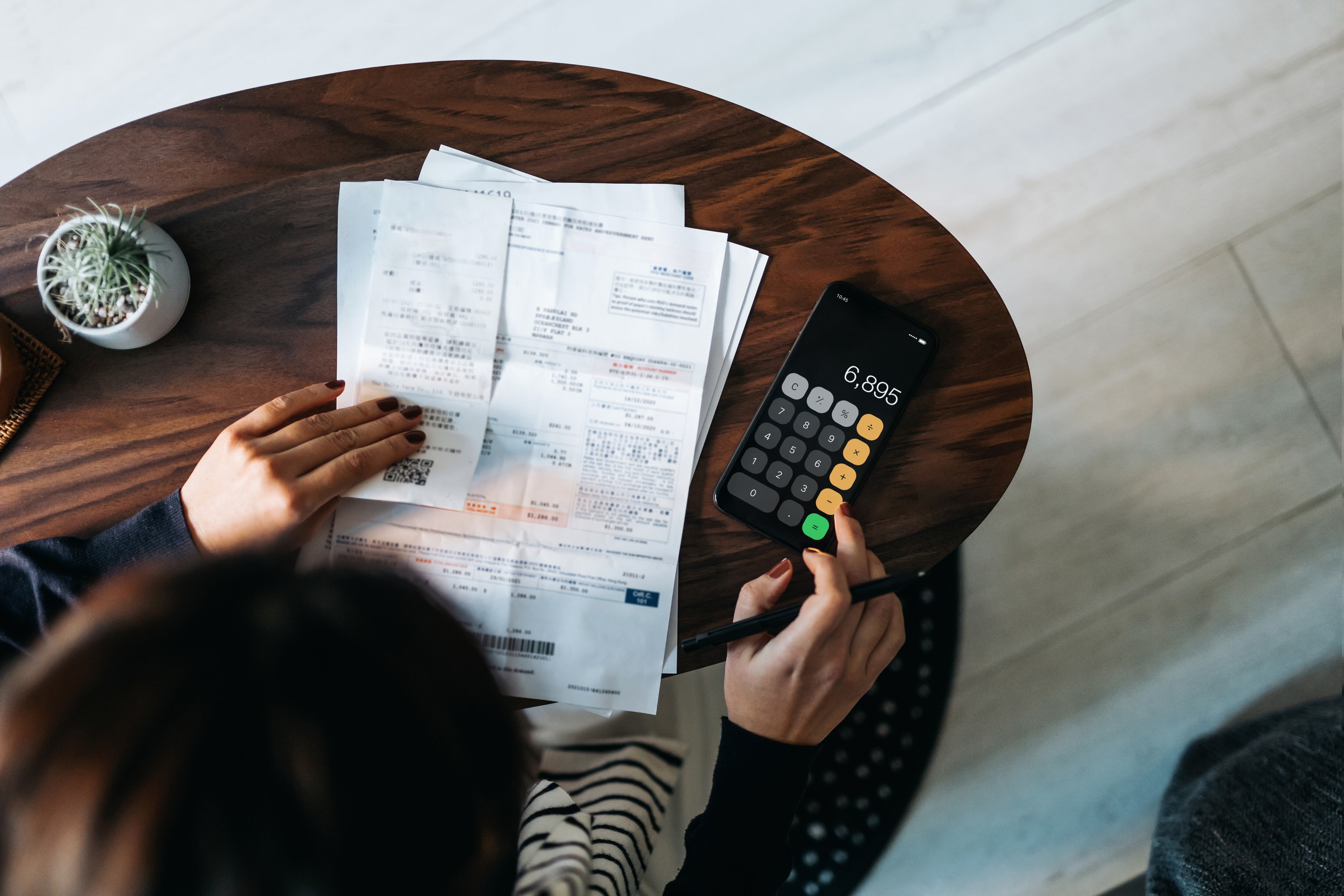 Overhead view of young Asian woman managing personal banking and finance at home. Planning budget and calculating expenses while checking her bills with calculator. Managing taxes and financial bills. Home budgeting. Concept of finance and economy