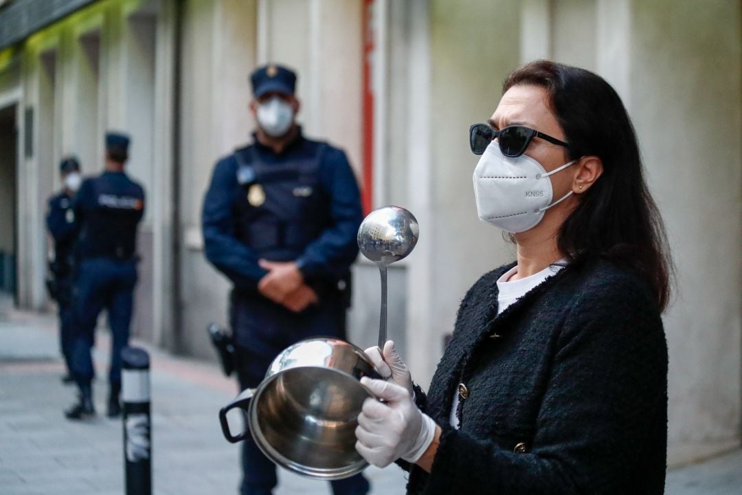 Una mujer con mascarilla golpea una cacerola delante de varios Policías Nacionales.