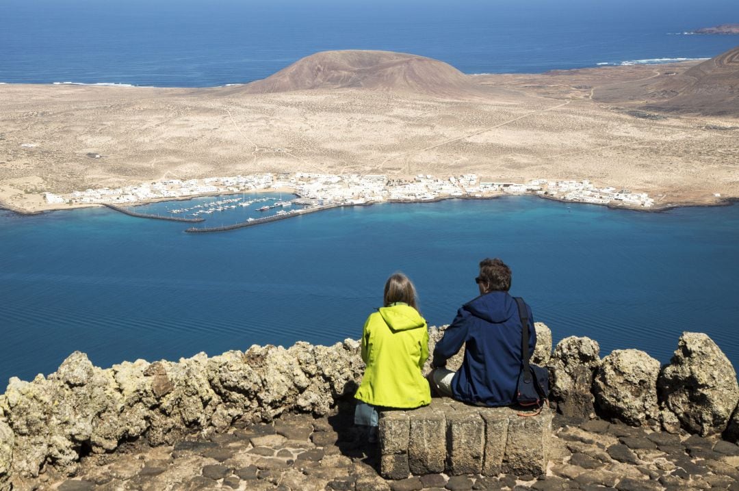 Dos turistas observan La Graciosa desde el Mirador del Río de César Manrique, al fondo, Caleta del Sebo