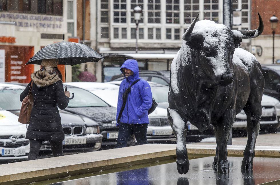 Dos personas caminan ante la escultura del toro ubicada en la conexión de la calle Santander con la avenida del Cid, en Burgos, que hoy amanecía bajo una intensa nevada.