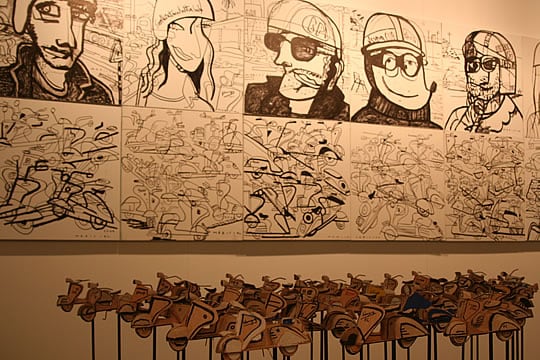 Imagen de una instalación que consiste de una serie de vespas de cartón suspendidas sobre un cable de acero y cuadros bitono estilo comic. Obra de Xavier Mariscal.