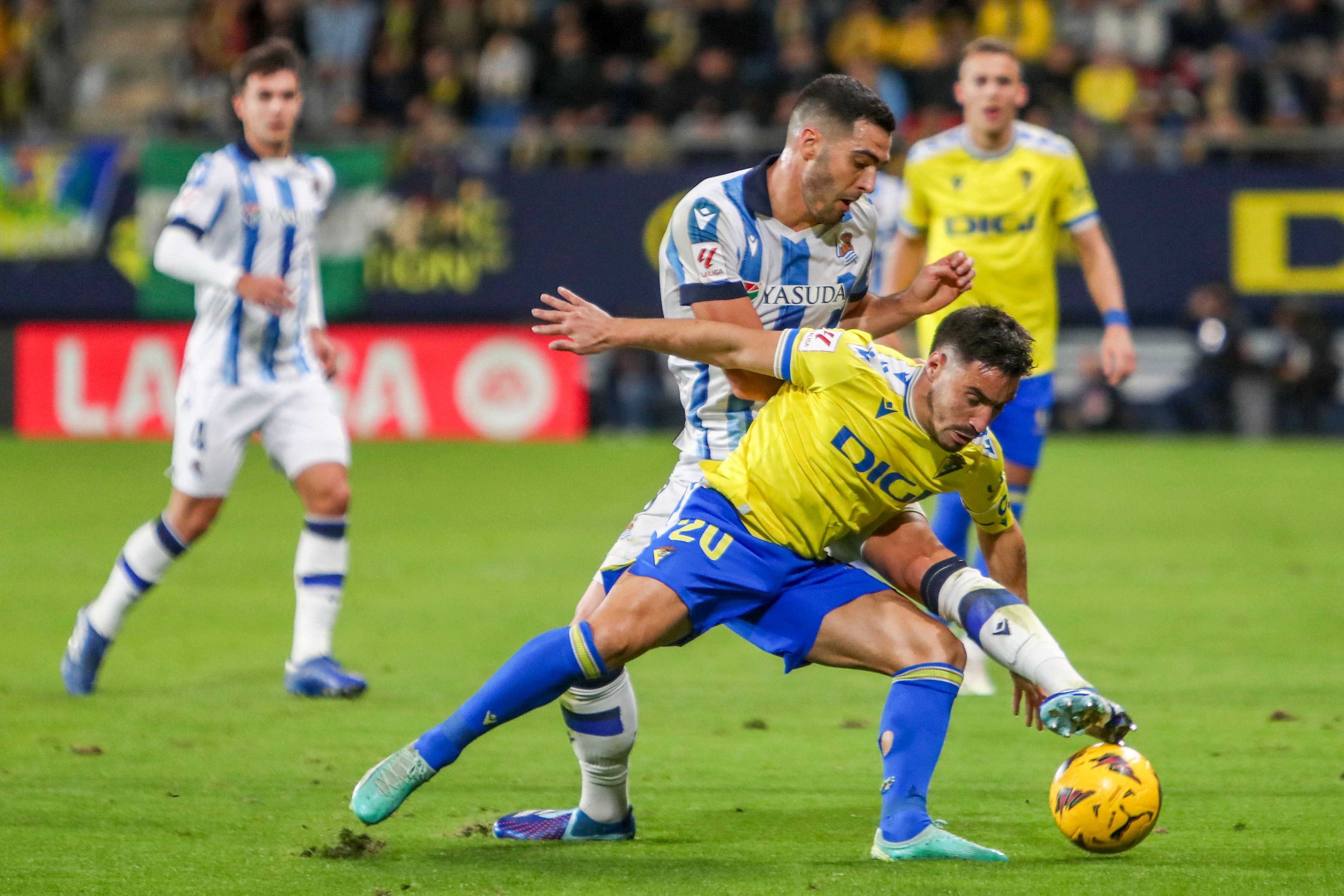 CÁDIZ, 21/12/2023. - El centrocampista del Real Sociedad Mikel Merino (detrás) lucha por el balón con el defensa del Cádiz CF Isaac Carcelén &quot;Iza&quot; durante el partido de LaLiga que enfrenta al Cádiz CF y la Real Sociedad este jueves en el Estadio Nuevo Mirandilla. EFE/Román Ríos.
