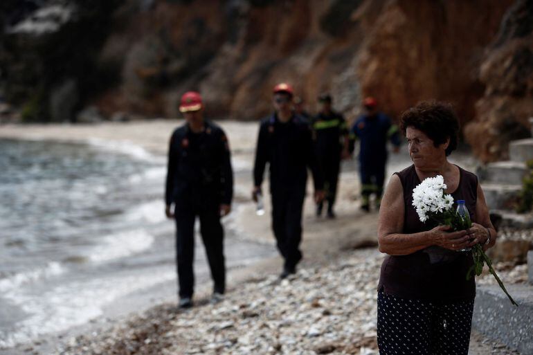 Varios bomberos (detrás) buscan a desaparecidos mientras una mujer sostiene un ramo de flores tras el incendio forestal de Mati, al noreste de Atenas (Grecia).