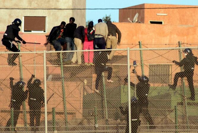 Varios inmigrantes africanos se sientan en la parte superior de la valla fronteriza, mientras agentes de la Guardia Civil española intentan evitar un que crucen a territorio español.