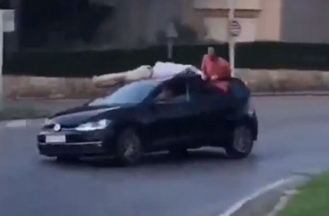 Un jove conduint un cotxe amb un noi estirat al sostre i un altre amb mig cos fora