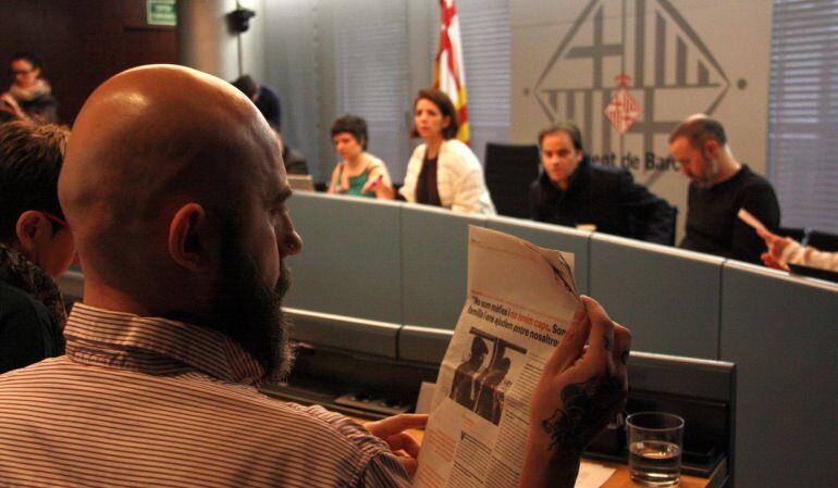 El regidor de la CUP, Josep Garganté, de perfil llegint un article de diari sobre manters que sosté amb la mà, amb els membres de la taula de la Comissió de presidència al fons