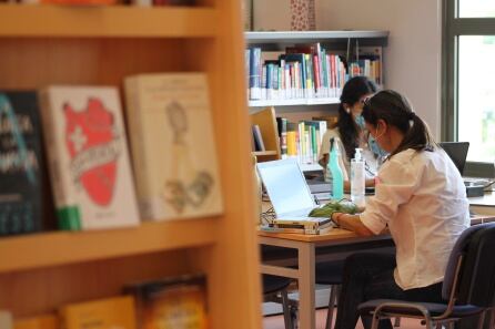 Dos trabajadoras en la biblioteca Miguel Delibes de Dos Hermanas. Ambas se encuentran sentadas en los extremos de la mesa, manteniendo la distancia, y llevan guantes y mascarilla.