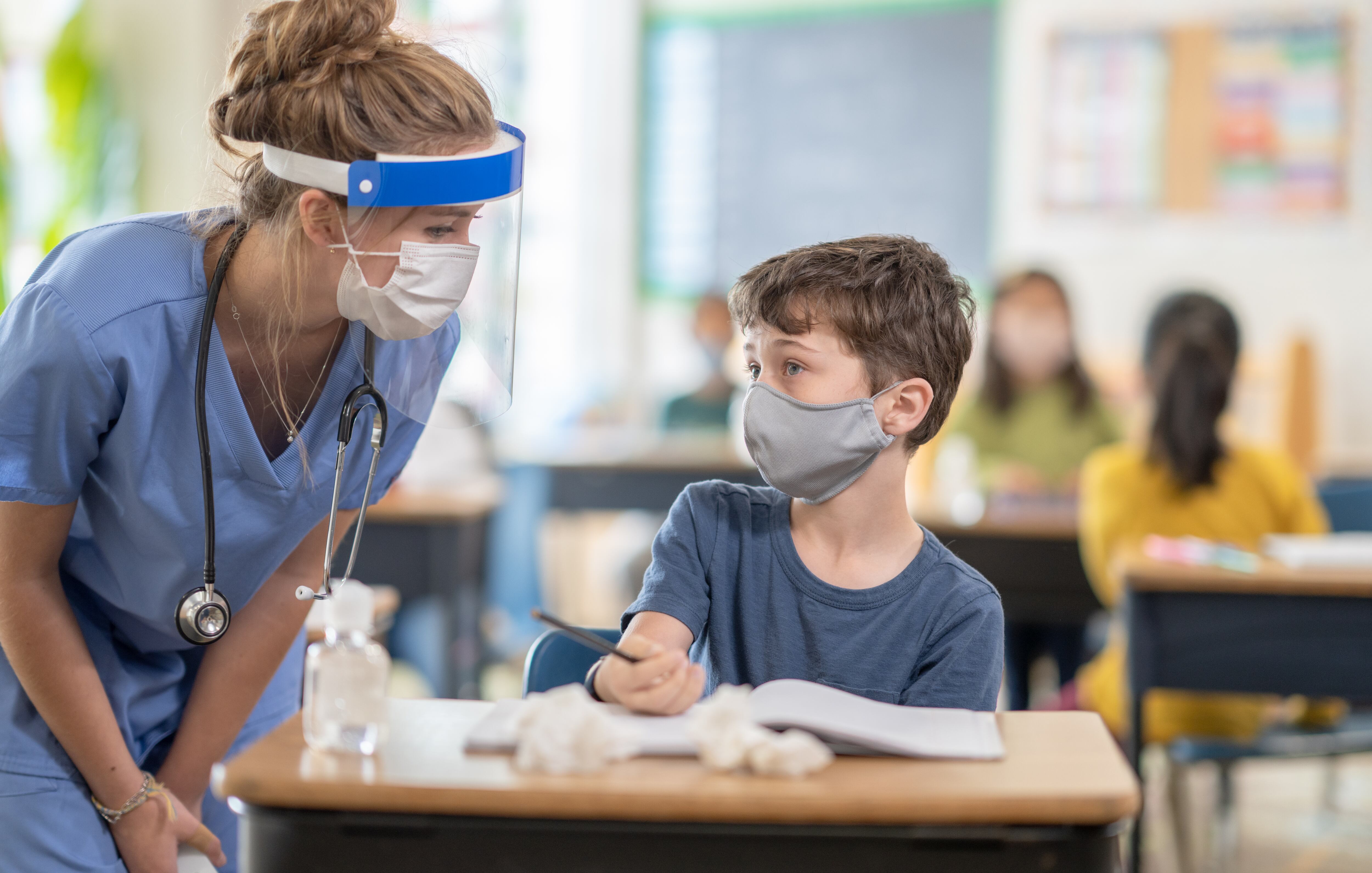 A female nurse is talking to an elementary school boy while he is sitting at his desk in a classroom.