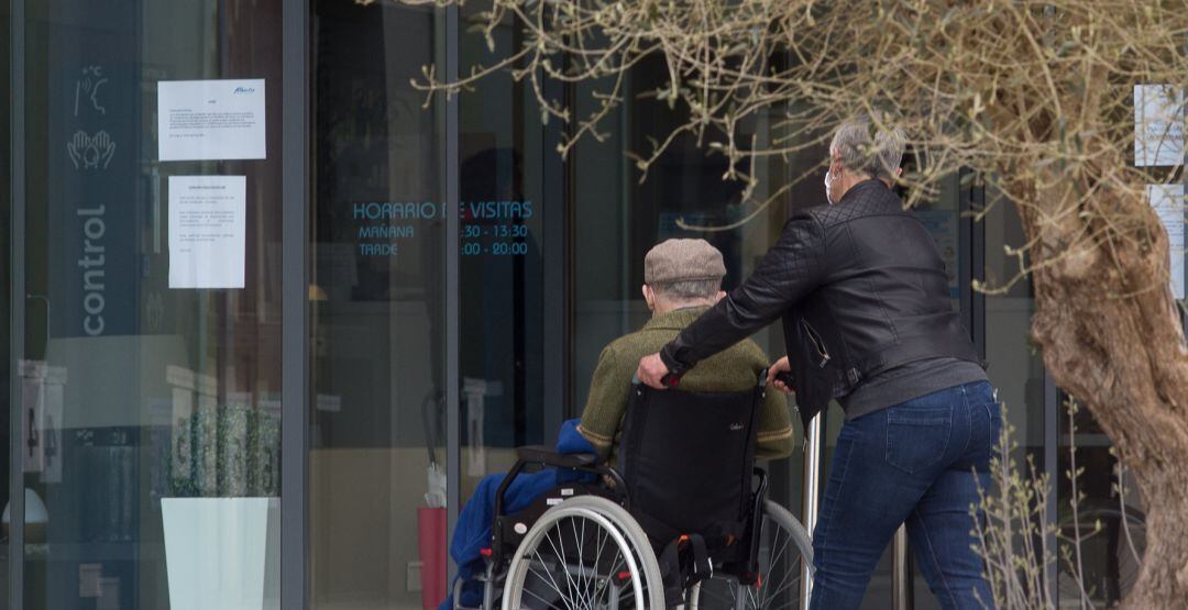 Una mujer pasea junto a un anciano en silla de ruedas hacia una residencia.
