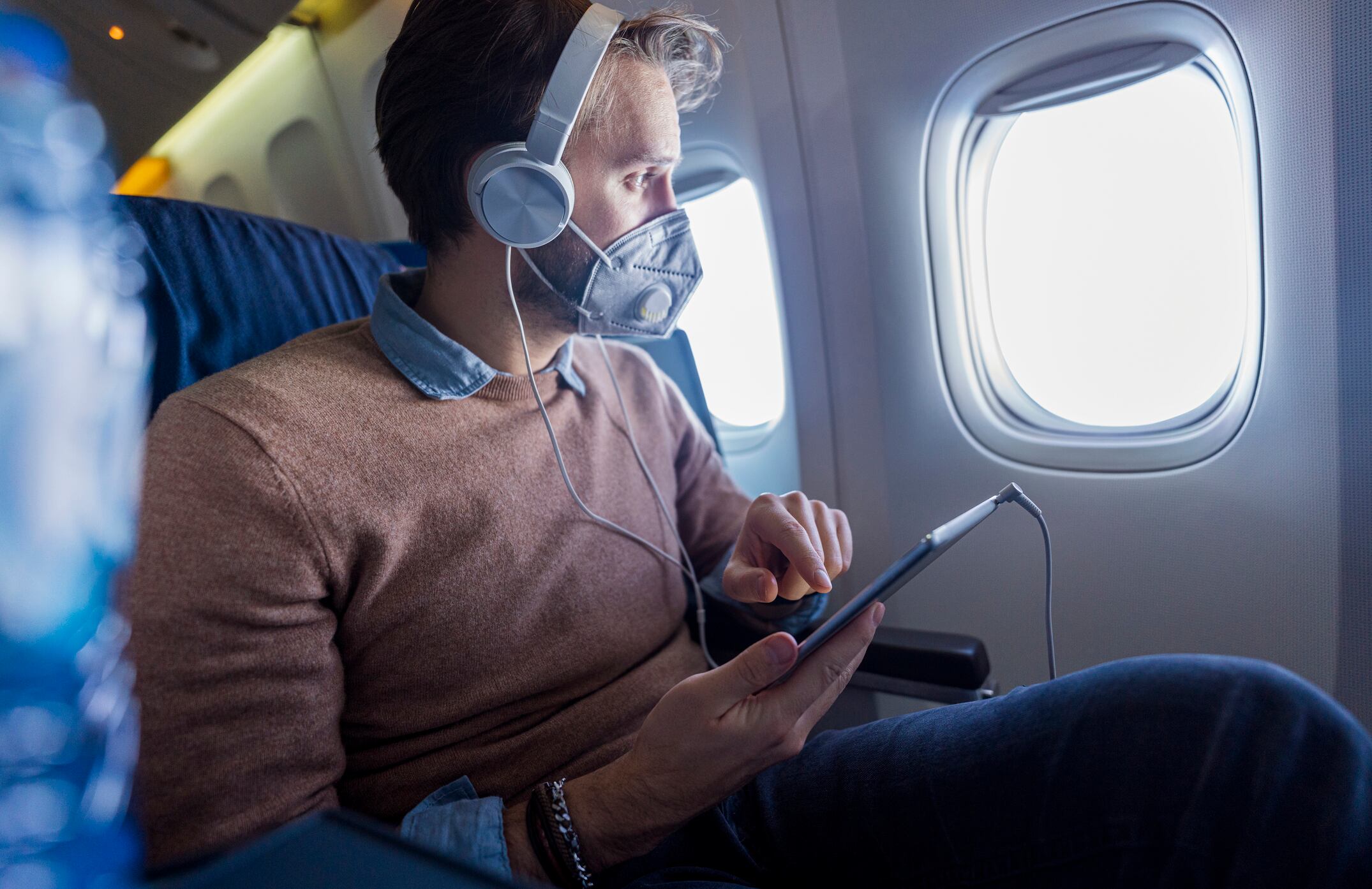 Young man in airplane seat listening music with tablet on his lap , he is wearing protection N95 mask during pandemic measures