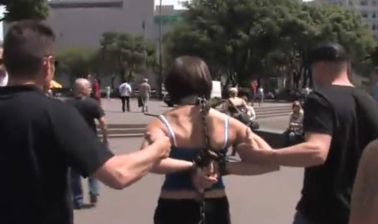 Una mujer encadenada es llevada por dos hombres en plena plaza Cataluña de Barcelona, a plena luz del día. Es una escena de vídeo porno rodado en la capital catalana