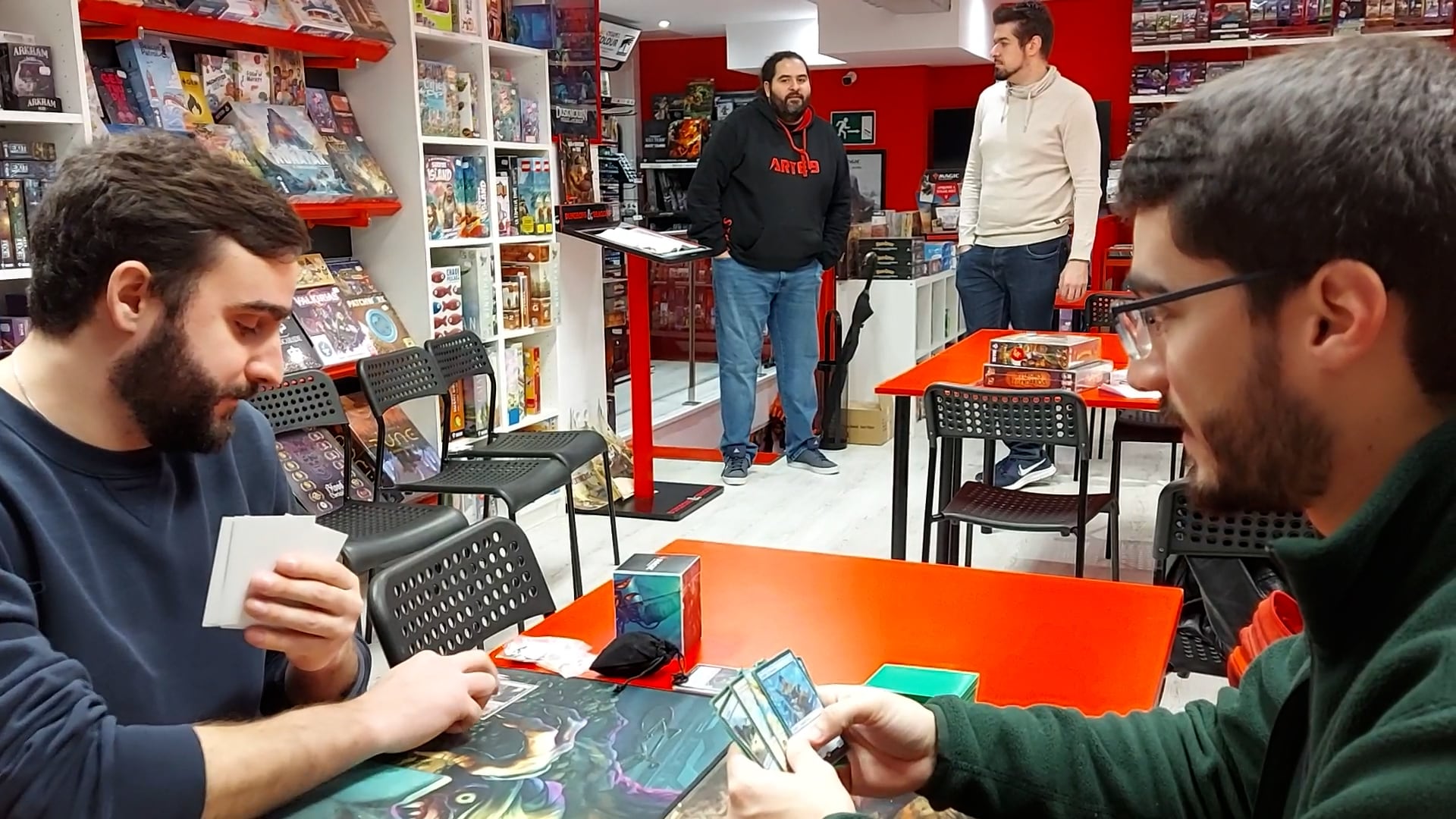 Dos aficionados juegan a Magic: The Gathering con el campeón del mundo al fondo