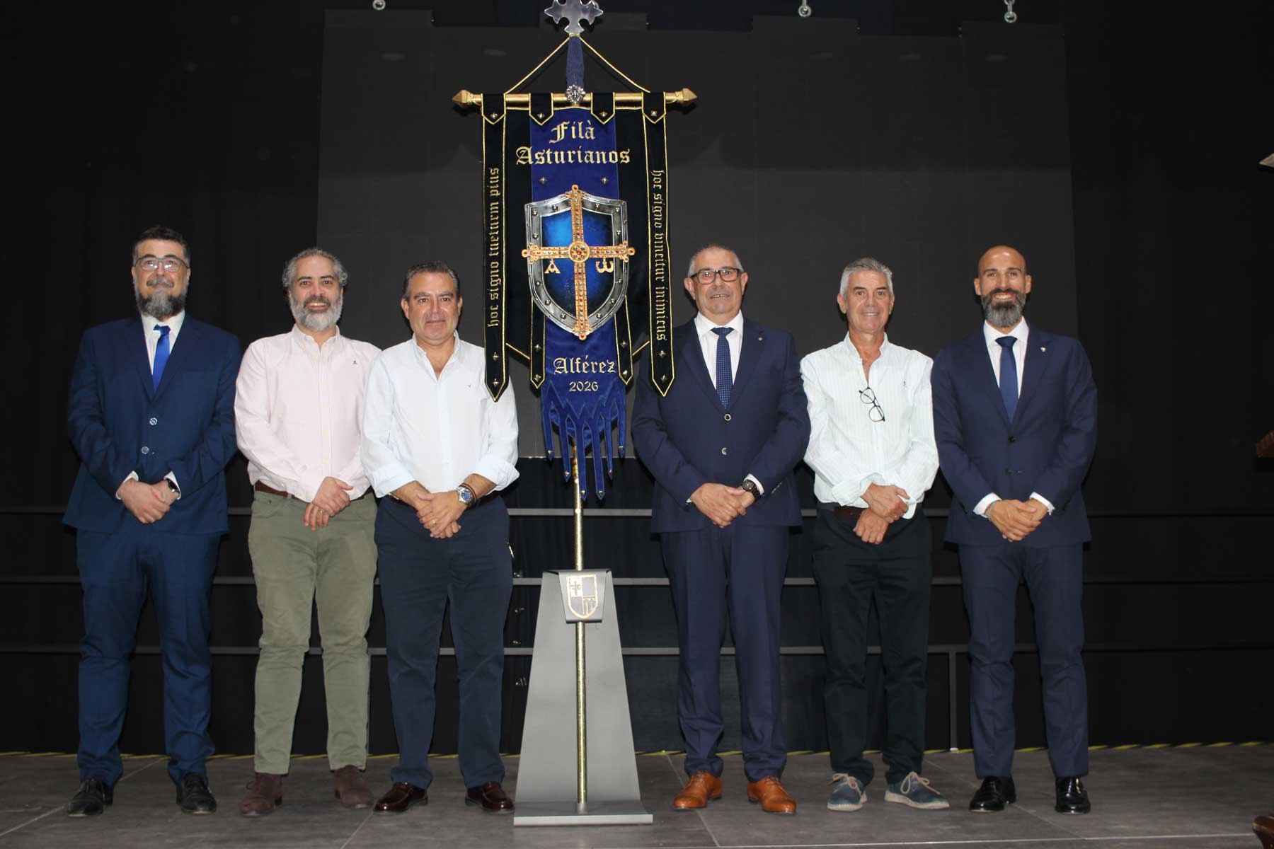 Los cargos del 2026, capitanes, alfereces y Mossén Torregrosa, junto al primer tro de los Asturianos, posan junto a la heráldica