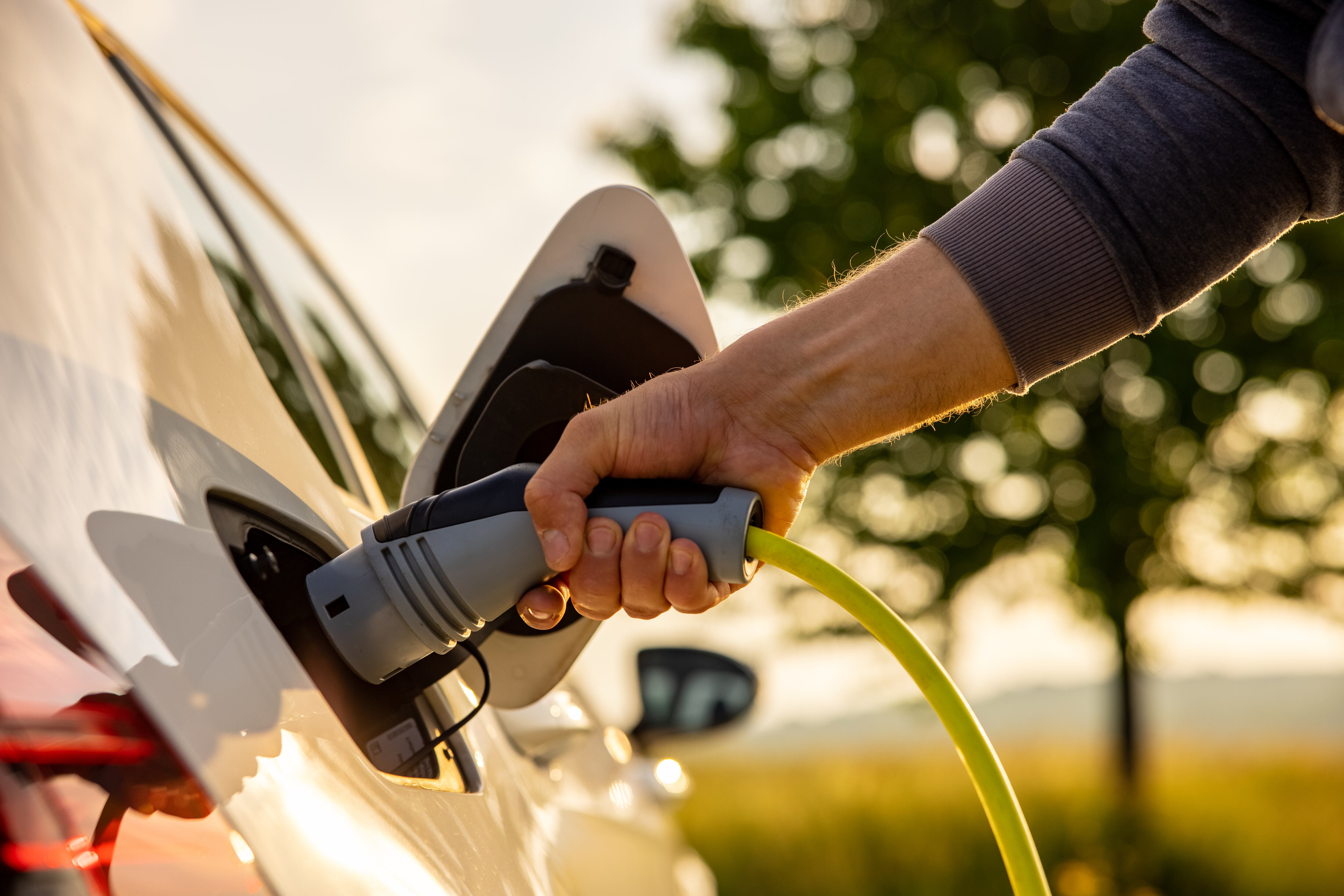 Hand of man inserting a power cord into an electric car for charging ecofriendly vehicle on green landscape