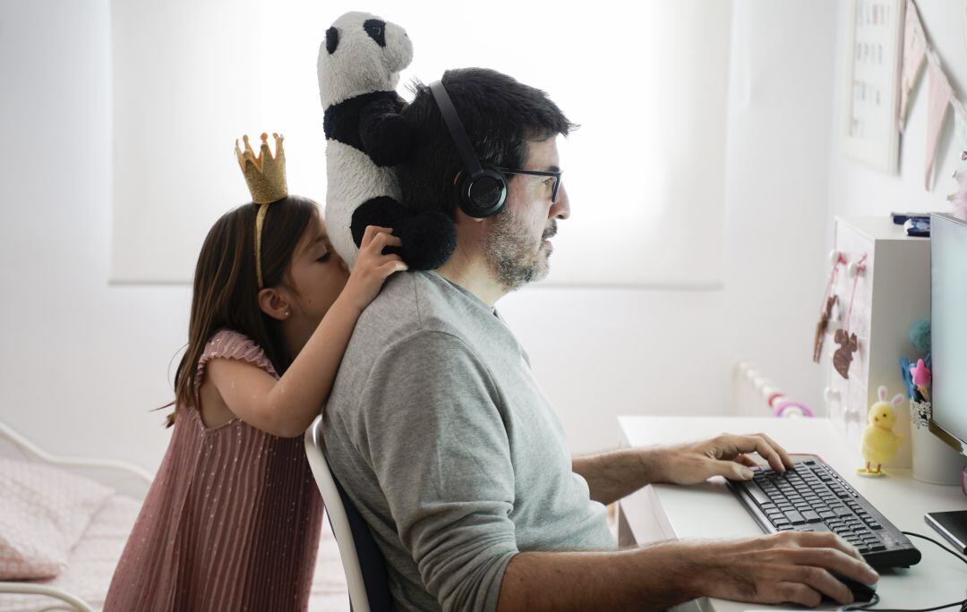 Padre teletrabajando en casa con su hija jugando cerca.