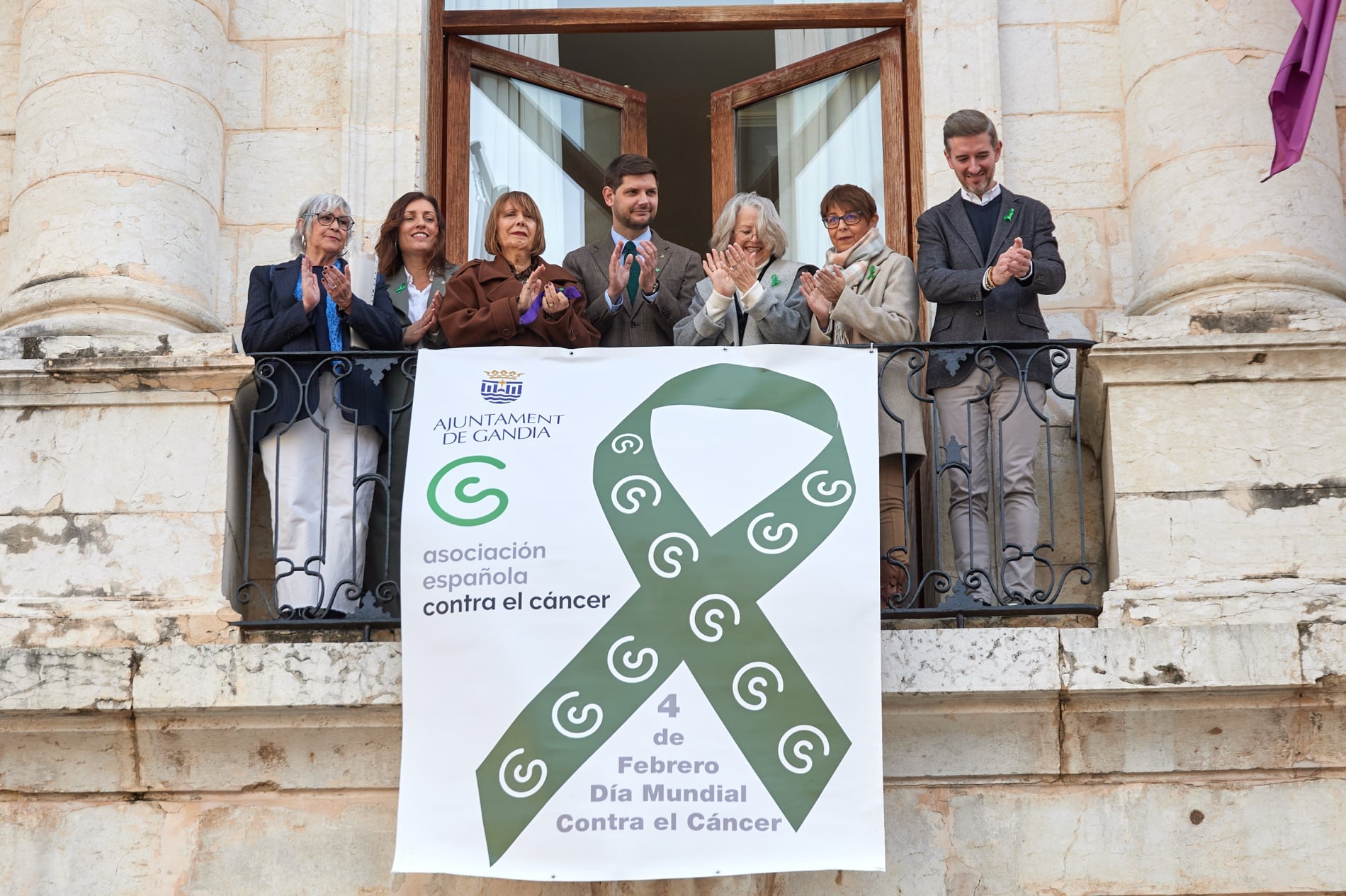Representantes de la Asociación Contra el Cáncer de Gandia y autoridades despliegan una pancarta en el balcón del Ayuntamiento.