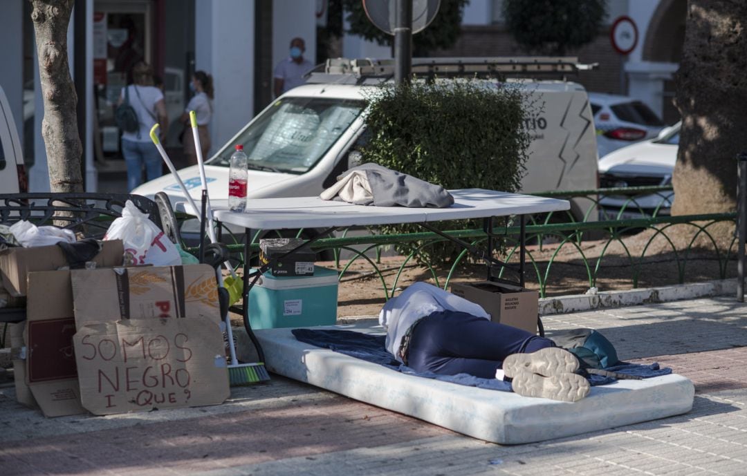 Entre pancartas, un hombre duerme sobre un colchón en plena calle, en la plaza donde se encuentra ubicado el ayuntamiento de Lepe (Huelva), donde en las últimas semanas dos incendios han calcinado los asentamientos precarios en los que vivían decenas de temporeros y temporeras migrantes que piden soluciones a su situación. 