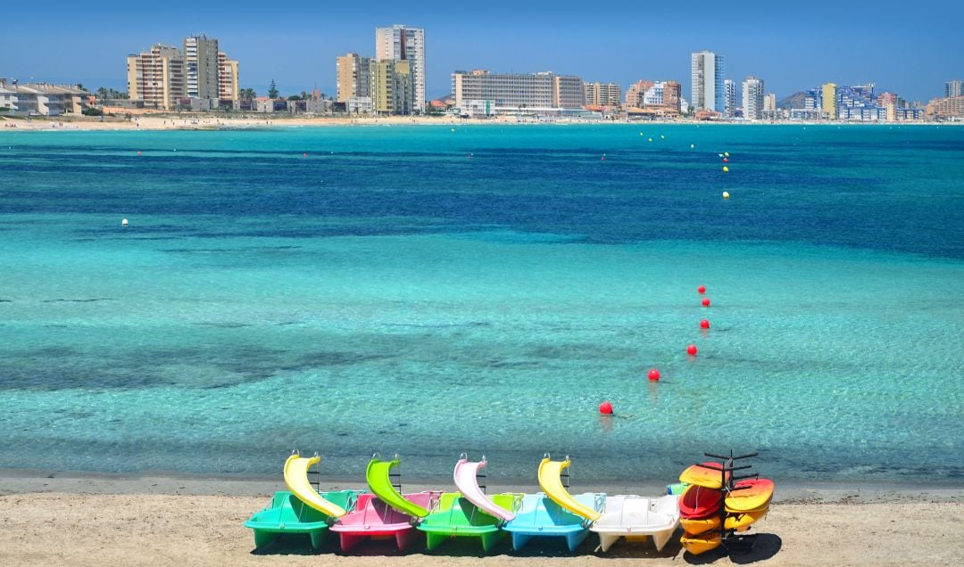 Un grupo de embarcaciones de colores amarradas en la arena con el fondo de las aguas turquesas de la playa de Levante, en verano, en Cabo de Palos, cerca de La Manga del Mar Menor, Murcia. Uno de los lugares más turísticos del levante español. Al fondo, el contraste de los grandes edificios de La Manga