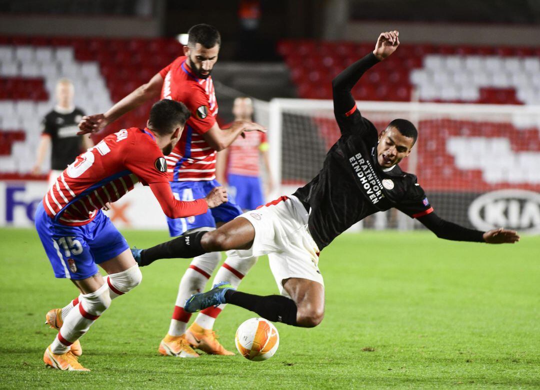 El delantero del PSV, Donyell Malen (d), cae ante los jugadores del Granada, Carlos Neva (i) y el francés, Maxime Gonalons, durante el encuentro correspondiente a la fase de grupos de la Liga Europa que han disputado este jueves en el estadio Nuevo Los Cármenes, en Granada.