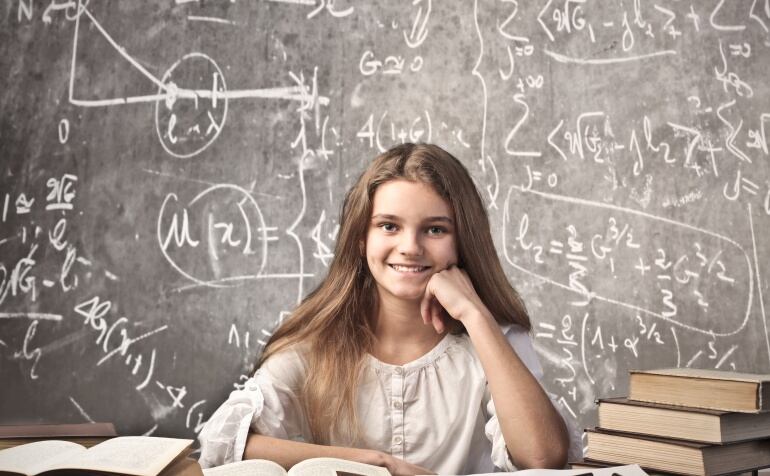 Imagen de una niña rodeada de libros delante de una pizarra llena de ecuaciones y fórmulas matemáticas.