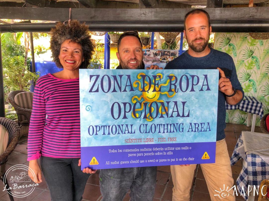 El presidente de la Asociación, Jesús Segura, la vicepresidenta, Ager Toro y el propietario del restaurante La Barraca, Isaac Rodriguezs muestran el cartel de &quot;Zona de ropa opcional&quot;