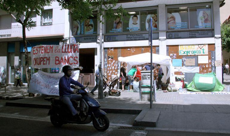 Una motocicleta circula per la Travessera de Gràcia passant just per davant de l'acampada instal·lada per el desallotjament del 'Banc Expropiat'