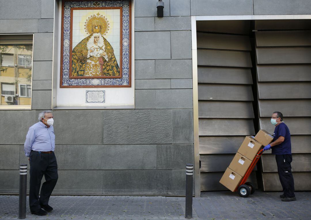 Un hombre con mascarilla pasea por Sevilla, cerca, otro hombre descarga cajas de material también debidamente protegido